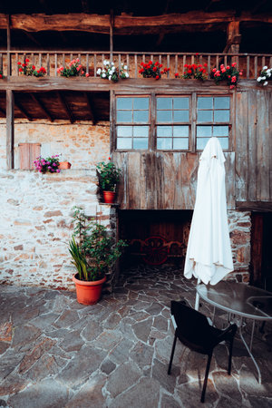 Rustic stone and timber farmhouse courtyard with wooden balcony, flower boxes, potted plants and patio table, closed white umbrella, vintage wagon wheelの写真素材