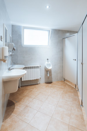 Wide-angle view of clean modern public restroom with tiled floor, wall-mounted sink, urinal, toilet stall partition, soap dispenser and bright window, minimal interior with copy spaceの写真素材