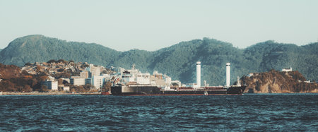 Telephoto view of a cargo ship fitted with Flettner rotor sails cruising past a coastal city and green mountains, calm blue sea and clear sky, copy space.の写真素材