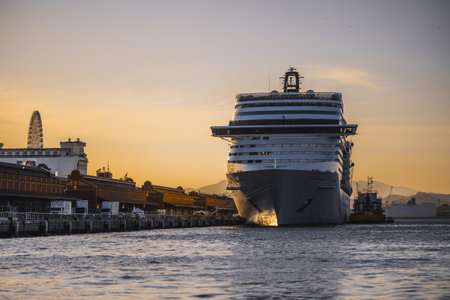 Huge cruise ship moored at harbor pier during golden sunset, front bow view near port terminal buildings, calm sea water, warm sky gradient and distant mountainsの写真素材