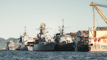 Telephoto view of gray naval warship and auxiliary vessel moored at dockyard pier, shipyard crane and warehouse buildings on shore, calm sea foreground, clear skyの写真素材