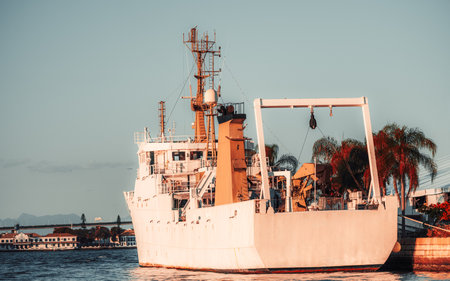 White industrial ship moored at harbor pier at sunset, stern view with crane gantry, mast antennas and bridge, calm water, palm trees and city coastlineの写真素材