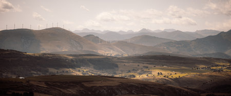 Panoramic view of rolling mountains and sunlit valleys with a line of wind turbines on distant ridges, patchwork fields, soft haze, and dramatic clouds at sunsetの写真素材