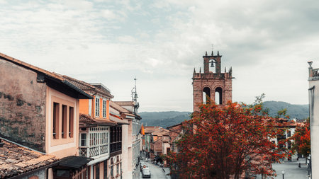 Autumn view of Salas, Asturias, Spain: colorful houses on a narrow street, historic stone church bell tower, red foliage tree, mountain backdrop, and dramatic cloudy skyの写真素材