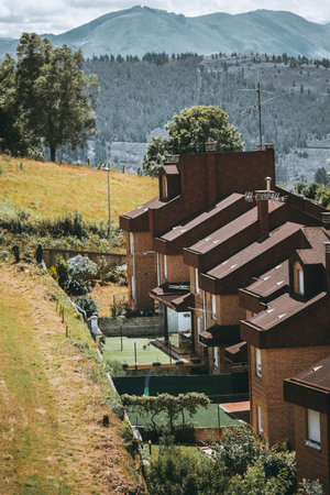 Telephoto view of a row of modern brick townhouses with brown tile roofs on a green hillside, fenced yards and small court, trees and dramatic mountain range in backgroundの写真素材