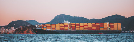 Telephoto view of large container ship fully loaded with colorful cargo containers sailing past coastal city and mountain landscape at dusk, calm sea foreground, global logistics and shippingの写真素材