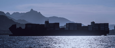 Backlit telephoto silhouette of large container ship on shimmering sea, stacked cargo box and faint smoke, with layered mountain ridge and hazy sky in dramatic blue hour lightの写真素材