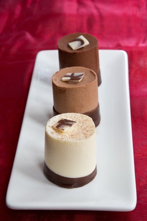 A trio of elegant chocolate mousses, comprising white chocolate, milk chocoalate & dark chocolate on a white plate against a red fabric backgroundの写真素材