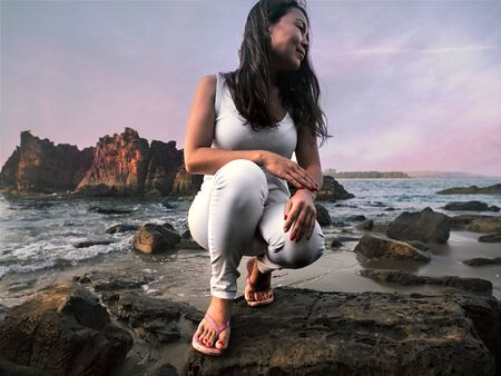 Asian girl sitting on the stone in front of the sea with rocksの写真素材