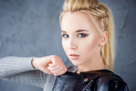 Gorgeous young woman with blond hair and perfect slim figure looking at camera wearing black leather jacket and gray dressの写真素材