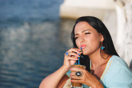 Happy tanned woman  drinking iced coffee in seaside cafe during sunset. Selective focus, copy spaceの写真素材