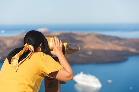 Beautiful young woman looking at cruise ships with binoculars in Thira, Santorini, Greece  enjoying...