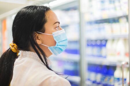 Woman buying food in supermarket. She is wearing protective mask and looking at the dairy section of a supermarket while shopping . Grocery shopping during COVID-19 concept. Selective focusの写真素材