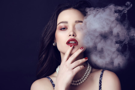 fashion studio photo of beautiful sensual woman with dark hair and bright makeup with bijou,posing in cigarette smokeの写真素材