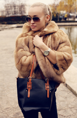 fashion outdoor photo of beautiful sensual girl with blond hair wearing luxurious fur coat and sunglasses, holding a bagの写真素材