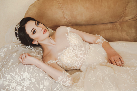 fashion photo of beautiful bride with dark hair in elegant wedding dress and diadem posing in room in the wedding morningの写真素材