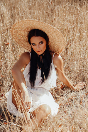 fashion summer outdoor photo of beautiful sexy woman with dark hair in elegant dress with luxurious straw hat and bag posing in field with dry grassの写真素材