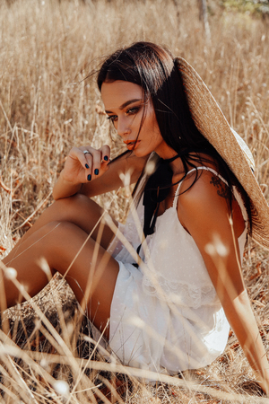fashion summer outdoor photo of beautiful sexy woman with dark hair in elegant dress with luxurious straw hat and bag posing in field with dry grassの写真素材