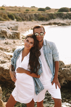 fashion outdoor photo of beautiful tender couple. gorgeous pregnant woman with dark hair in elegant dress posing with her handsome husband on the summer beachの写真素材