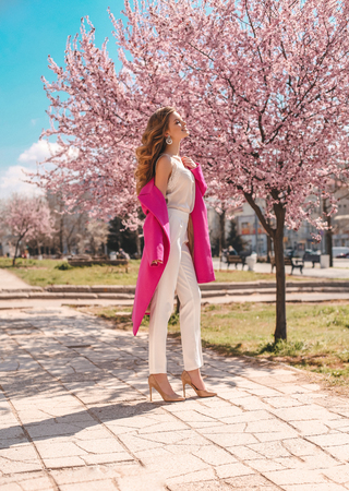 fashion outdoor photo of beautiful young girl with natural hair color in elegant clothes and pink coat posing among flowering peach trees in spring gardenの写真素材