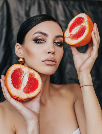 fashion studio photo of beautiful woman with dark hair with evening makeup posing with grapefruitの写真素材