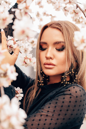 fashion outdoor photo of beautiful young girl with blond hair in elegant clothes and earrings posing among flowering peach trees in spring gardenの写真素材