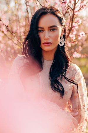 fashion outdoor photo of beautiful sensual woman with dark hair in elegant dress posing among flowering peach trees in gardenの写真素材