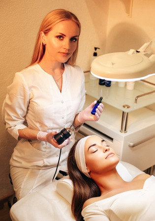 fashion photo of beautiful woman with dark hair and glowing skin making ultrasound therapy procedure in beauty salonの写真素材