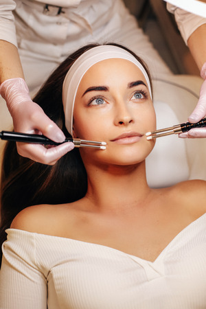 fashion photo of beautiful woman with dark hair and glowing skin making micro-current therapy in beauty salonの写真素材