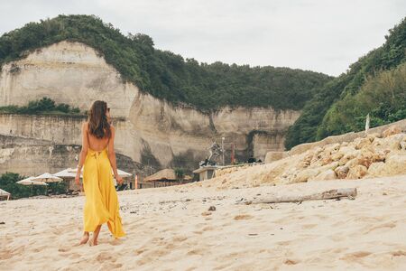 Asia travel series. Beautiful woman with dark hair in elegant yellow dress walking on the sand in Balinese beachの写真素材