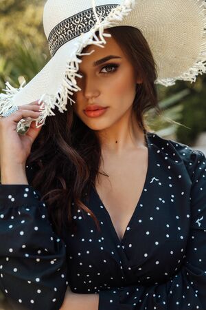 fashion outdoor photo of beautiful woman with dark hair in elegant dress and hat posing in summer garden