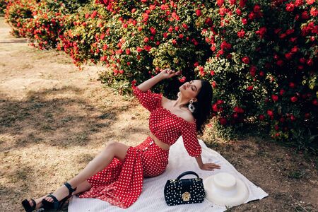 fashion outdoor photo of beautiful sensual woman with dark hair in luxurious dress and accessories posing in summer garden with red rosesの写真素材