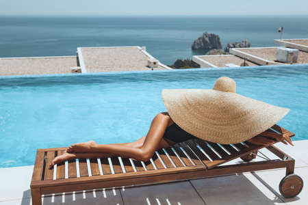 fashion outdoor photo of beautiful sexy woman with blond hair in bikini and big straw hat posing near swimming pool with sea viewの写真素材