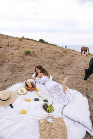 fashion outdoor photo of beautiful woman with blond hair in elegant dress having picnic with mountains view and horses on backgroundの写真素材