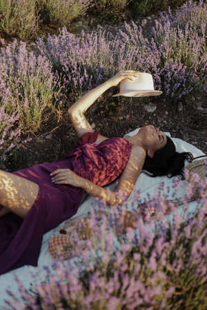fashion outdoor photo of beautiful woman with dark hair in elegant dress with accessories having picnic in blooming lavender fieldの写真素材