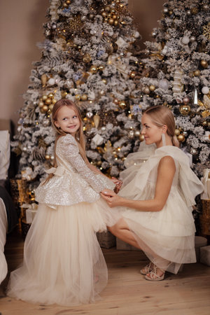 Fashion photo of beautiful family, mother and her daughter with blond hair in elegant dresses posing in decorated studio with Christmas tree and presentsの写真素材