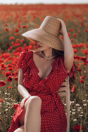 fashion outdoor photo of beautiful woman with dark hair in elegant red dress and straw hat posing in blooming poppy fieldの写真素材