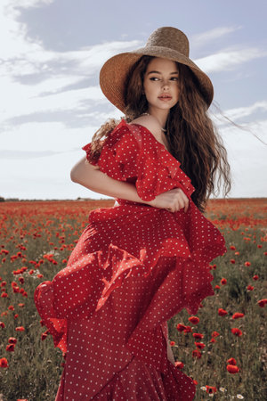 fashion outdoor photo of beautiful woman with dark hair in elegant red dress and straw hat posing in blooming poppy fieldの写真素材