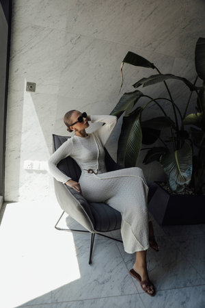 fashion photo of beautiful woman in elegant white dress with accessories posing in the room sitting on sofaの写真素材
