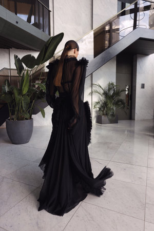 fashion photo of beautiful woman with dark hair in elegant black dress with accessories posing in luxurious hotel lobbyの写真素材