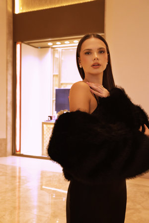 fashion interior photo of beautiful sensual woman with dark hair in luxurious black fur coat and jewelry posing in hall of the hotelの写真素材