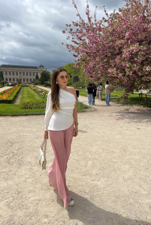 fashion outdoor photo of beautiful woman with dark hair in elegant clothes with accessories posing in spring garden with blossom sakura treeの写真素材