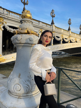 fashion outdoor photo of beautiful woman with dark hair in luxurious white silk blouse and black pants and accessories posing by the Alexandre III bridge in Parisの写真素材