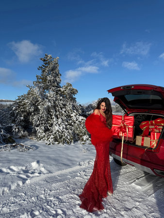 fashion Christmas photo of beautiful woman with dark hair in luxurious evening dress with accessories posing in the snowy mountains by a car with a trunk full of gift boxesの写真素材