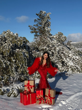 fashion Christmas photo of beautiful woman with dark hair in luxurious evening dress with accessories posing in the snowy mountains with a red gift box with bowの写真素材