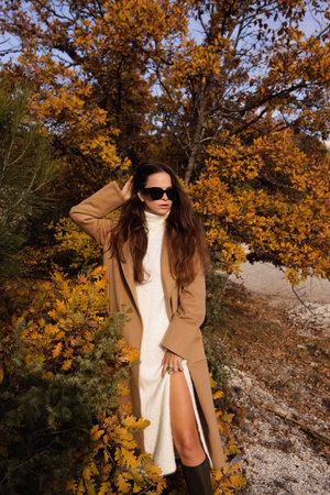 fashion outdoor photo of beautiful woman with dark hair in elegant dress and wool coat posing among beautiful natureの写真素材