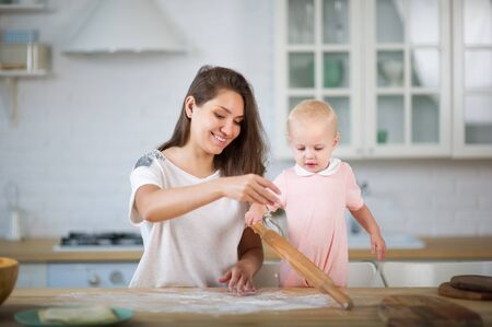 a young mother in the bright kitchen plays with her daughter, sprinkling flour on the countertop, and the daughter at this time plays with a rolling pin and looks at the falling flourの写真素材