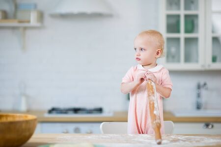 little blue-eyed blonde girl stands near the countertop and holds a rolling pin in his handの写真素材