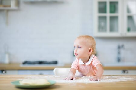 little blue-eyed blonde girl leads her hands on the countertop with flour and looks awayの写真素材