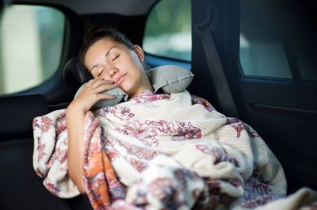 the girl on the road is tired and sleeps in the back seat in the car, hiding in a blanketの写真素材